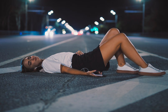 Woman in earbuds lying on night road listening to music in earphones