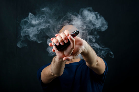 hand holding vape e-cigarette or electronic cigarette with white smoke over a black background. 