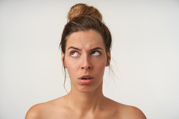 Portrait of attractive young woman wearing bun hairstyle and no make-up, looking upwards with bored face, standing over white background