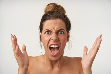 Close-up of wrathful young lovely woman posing over white background with raised palms, frowning and shouting with wide mouth opened