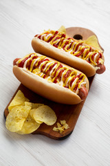 Homemade colombian hotdogs with pineapple sauce, chips, yellow mustard and mayo ketchup on a rustic wooden board on a white wooden table, side view.
