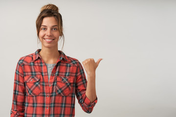 Pretty lady with charming smile showing thumb aside, wearing checkered shirt and natural make-up, looking cheerfully to camera, standing over white background