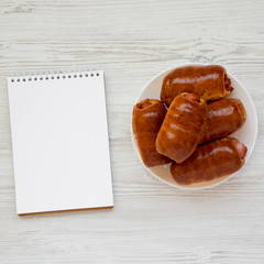 Homemade beef sausage kolache on a white plate, blank notepad on a white wooden surface, top view. Copy space.
