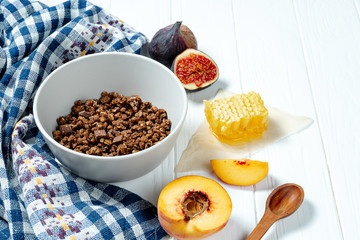 Chocolate granola in a white bowl in a composition with honeycombs, a spoon, figs and peach. Healthy breakfast food. Making breakfast. Gluten free