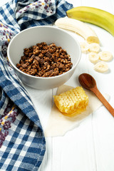 Chocolate granola in a white bowl in a composition with honeycombs, a spoon, banana on white wooden background. Healthy breakfast food. Making breakfast. Gluten free