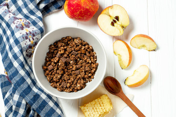 Chocolate granola in a white bowl in a composition with honeycombs, a spoon, apple on white wooden background. Healthy breakfast food. Making breakfast. Gluten free