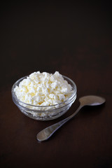 Cottage cheese crumbly in a glass transparent bowl and metal spoon on a wooden old scratched brown tabletop.