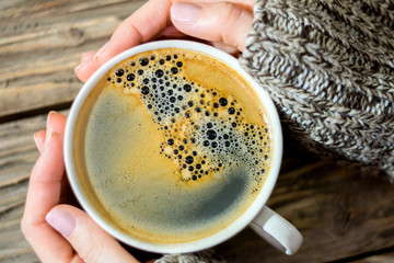 A cup of fragrant hot coffee with foam, held by female hands in a knitted sweater against the background of an old textured wooden table