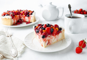 A piece of home-made cottage cheese berry pie. Garnished with cream, strawberries, blueberries, sea buckthorn berries. On a white plate. Next to the plate is a fork, a napkin, strawberries. 