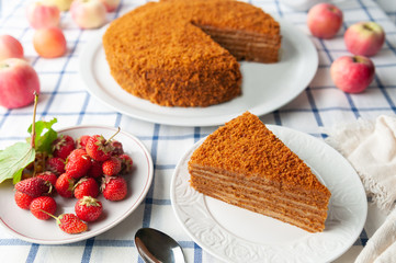 A slice of homemade multilayer honey cake. Orange color. On a white plate. Nearby are berries, apples and cake. Tablecloth in a rustic style.
