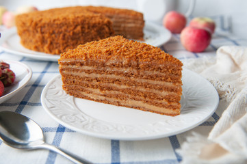 A slice of homemade multilayer honey cake. Orange color. On a white plate. Close-up. Testura. Nearby are berries, apples and cake. Tablecloth in a rustic style.