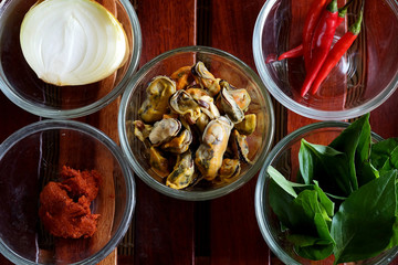 Set of important ingredients for making Stir Fried Mussels with Thai Basil and Chili on wood mat and space for write wording containing Mussels, Onion, Red Chili Paste, Chili and Thai Basil leaves