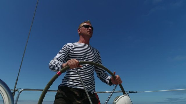 A young man sailor at the helm of a sailing yacht turns the steering wheel, controls the movement and looks forward putting his hand covering the sun.