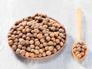 Allspice (Jamaica pepper) in clay plate and wooden spoon vertically on grey cement background
