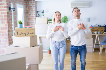 Fototapeta na wymiar Young beautiful couple standing at new home around cardboard boxes smiling with hands on chest with closed eyes and grateful gesture on face. Health concept.