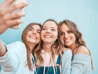 Three young smiling hipster women in summer clothes.Girls taking selfie self portrait photos on smartphone.Models posing in the street near wall.Female showing positive face emotions