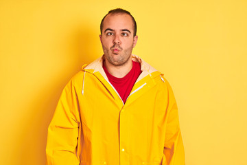 Young man wearing rain coat standing over isolated yellow background making fish face with lips, crazy and comical gesture. Funny expression.