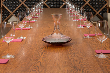 on a large wooden table there are a lot of wine glasses and a decanter with wine, tasting is expected, in the background shelving