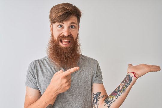 Young redhead irish man wearing t-shirt standing over isolated grey background amazed and smiling to the camera while presenting with hand and pointing with finger.