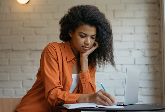 Attractive university student using laptop computer, studying, learning language, exam preparation at library. Pensive African American woman freelancer writes notes, planning working process