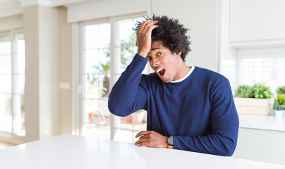 Young african american man wearing casual sweater sitting at home surprised with hand on head for...