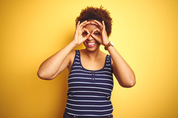 Beauitul african american woman wearing summer t-shirt over isolated yellow background doing ok gesture like binoculars sticking tongue out, eyes looking through fingers. Crazy expression.