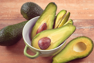 Close-up photo of fresh sliced avocados, brown seeds visible in green bowl on brown wooden background. Vegetarian food concept, vintage style.