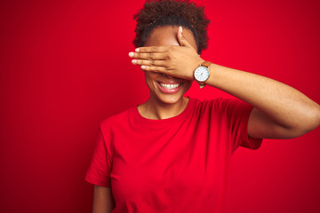 Young beautiful african american woman with afro hair over isolated red background smiling and laughing with hand on face covering eyes for surprise. Blind concept.