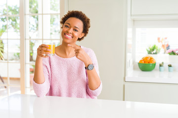 Young african american woman driking orange juice at home very happy pointing with hand and finger