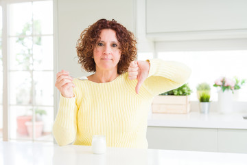 Senior woman eating a healthy natural yogurt at home with angry face, negative sign showing dislike with thumbs down, rejection concept