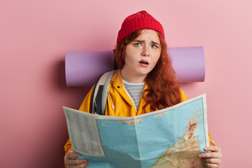 young puzzled ginger beautiful woman wearing care mat, knitted cap, backpack, yellow jacket, shirt...