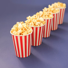 flavored crispy popcorn poured into five striped white-red paper cups, which stand in a row diagonally on a lilac background, copy space, close-up, square