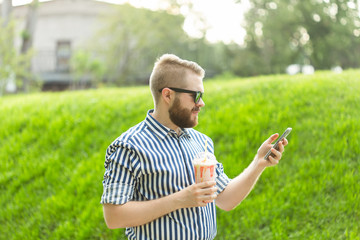 Naklejka na ściany i meble Positive young male hipster listens to your favorite online radio using smartphone headphones and high-speed wireless internet in the park on a sunny warm summer day.
