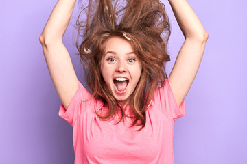 Close up portrait shot of playful young female jumping high, shouting loudly, messy hair upwards, suprised and happy to get present, dreams come true, birthday girl, modeling over light purple wall.