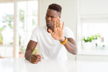 Handsome african american man on white table at home doing stop sing with palm of the hand. Warning expression with negative and serious gesture on the face.