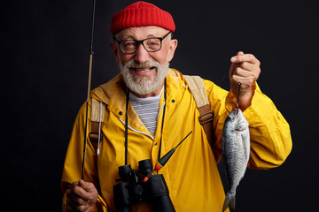 old experienced happy fisherman in glasses holding a fish and a fishing rod isolated on black background. close up photo. studio shot. hobby, interets