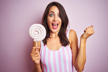 Young beautiful woman eating sweet candy over pink isolated background screaming proud and celebrating victory and success very excited, cheering emotion