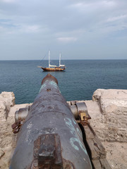 The old large caliber cannon aimed at the sailing ship on the sea.