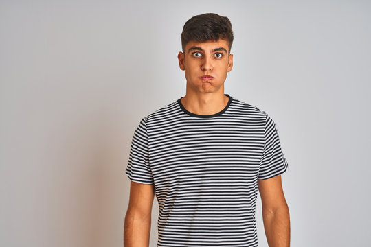 Young indian man wearing navy striped t-shirt standing over isolated white background puffing cheeks with funny face. Mouth inflated with air, crazy expression.