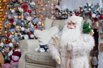 Santa Claus in glasses dressed in a white fur coat, holding wooden skis on the background of rustic chairs, plush toys husky Christmas tree with lights garland