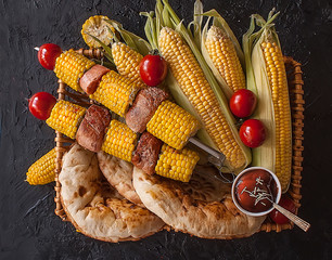 Obraz na płótnie Canvas Shish kebab of corn and meat and tortilla in a basket on a black dark background. Fried vegetables. Corn, tomato, pepper. Roasted meat. Grilled vegetables. Top view