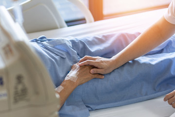 Caregiver holding elderly senior patient (ageing old adult person) hand in hospital bed or nursing hospice, geriatrician palliative home, while caretaker having  medical health care service