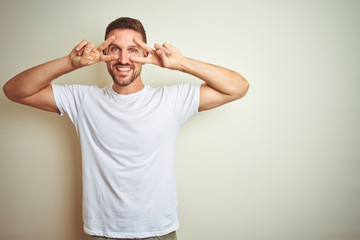 Young handsome man wearing casual white t-shirt over isolated background Doing peace symbol with fingers over face, smiling cheerful showing victory