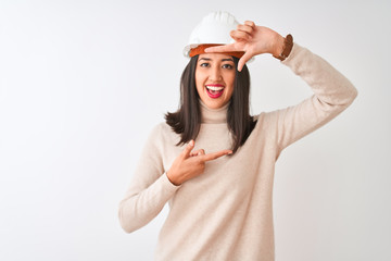 Young chinese architect woman wearing security helmet over isolated white background smiling making frame with hands and fingers with happy face. Creativity and photography concept.