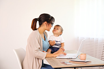 Stay at home mom working remotely on laptop while taking care of her baby. Young mother on maternity leave trying to freelance by the desk with toddler child. Close up, copy space, background.