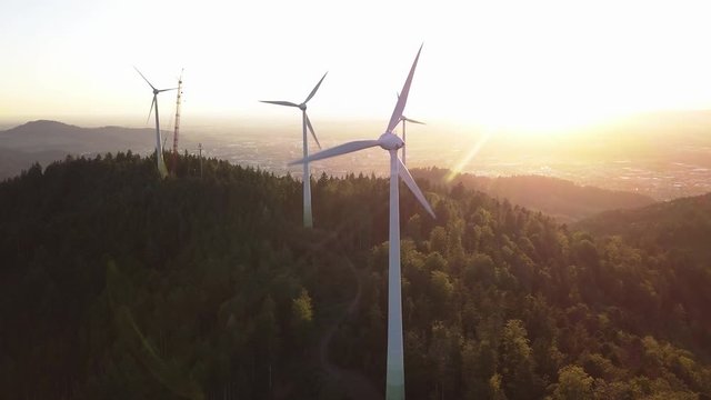 Orbiting aerial view of wind turbines in the hills during sunset with mountains in the background. Propellers tickling the sun while a crane stands at an inactive turbine