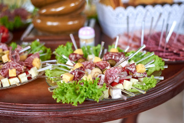 Appetizing sausage, cheese, lettuce, tomatoes, olives are put on skewers laid out on a plate on a table.