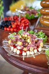 Appetizing sausage, cheese, tomatoes, olives are put on skewers laid out on a plate on a table.