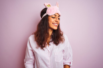 Young beautiful woman wearing pajama and sleep mask over isolated pink background looking away to side with smile on face, natural expression. Laughing confident.