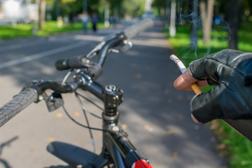 cigarette in the hand of a smoking cyclist who rides a bike on the background of the asphalted path of the city park